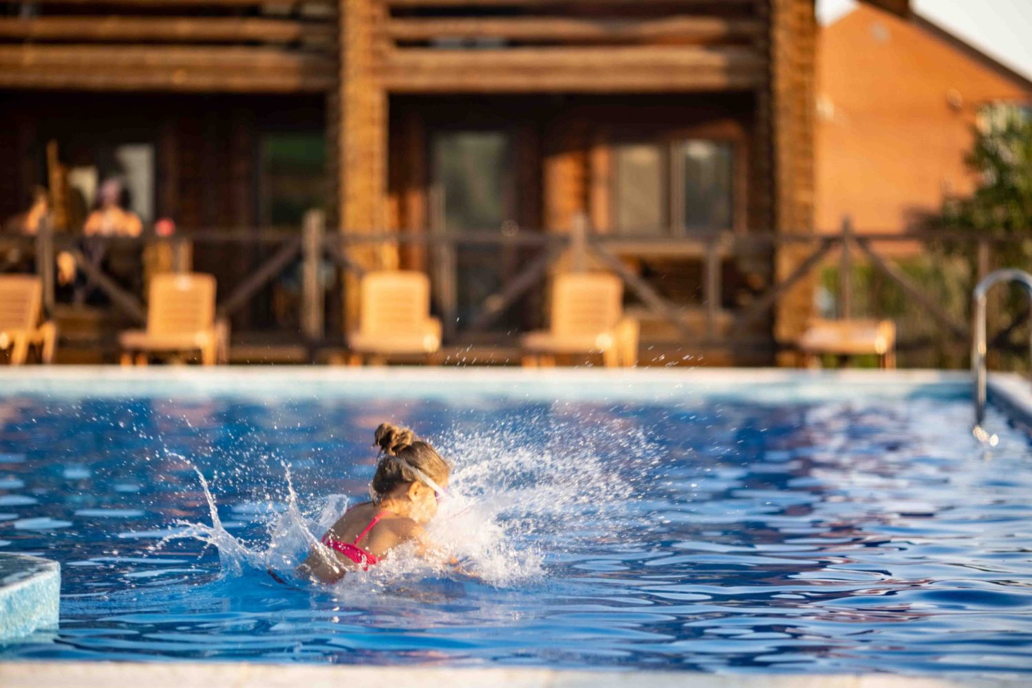 A girl in bright pink swimsuit and black swimming goggles, funny jumping into deep blue pool with clean transparent cool water against background of the warm summer sunny sunset