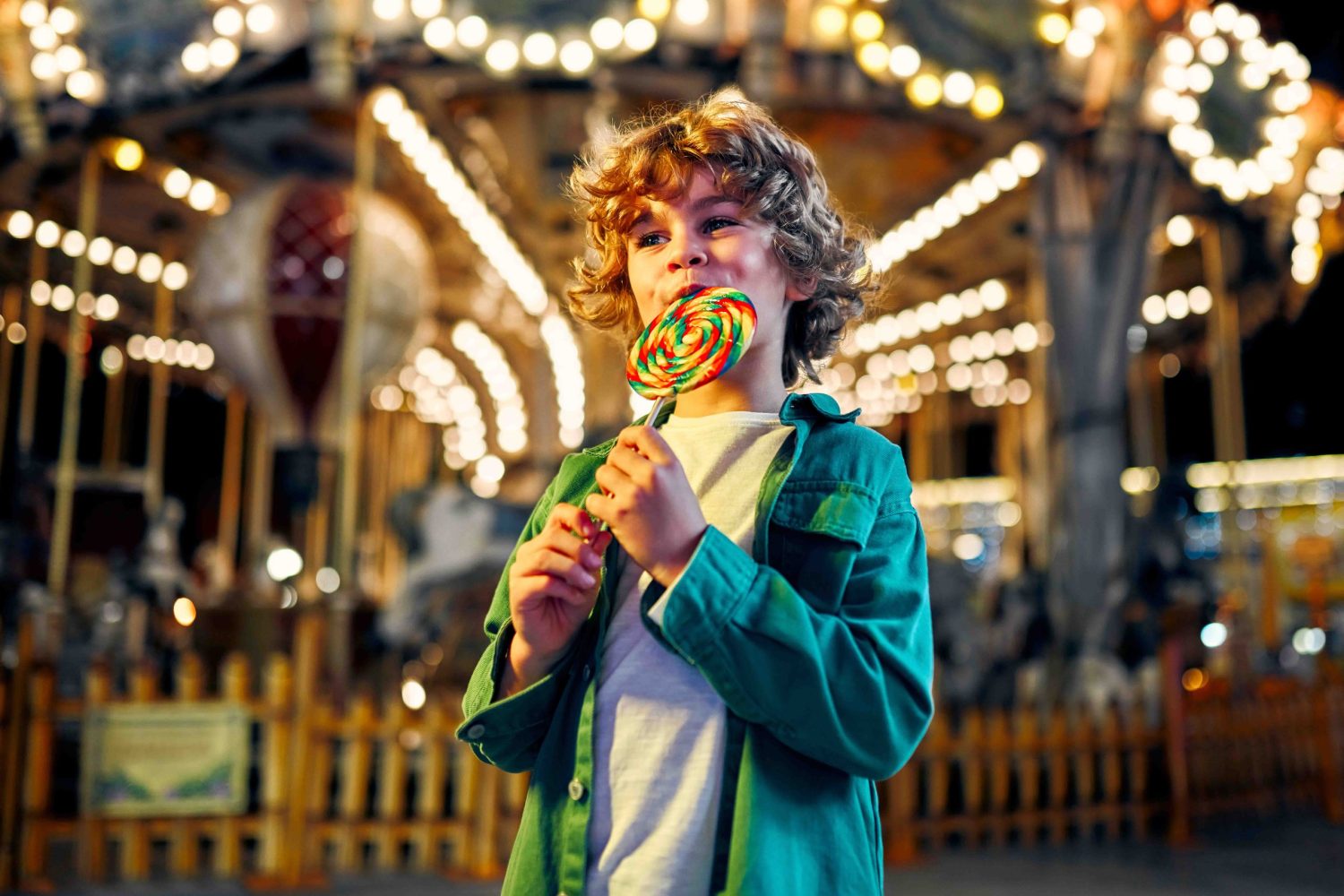 A cute caucasian boy with blonde curly hair eating a colorful lollipop standing against the background of a carousel with horses in the evening at an amusement park or circus.